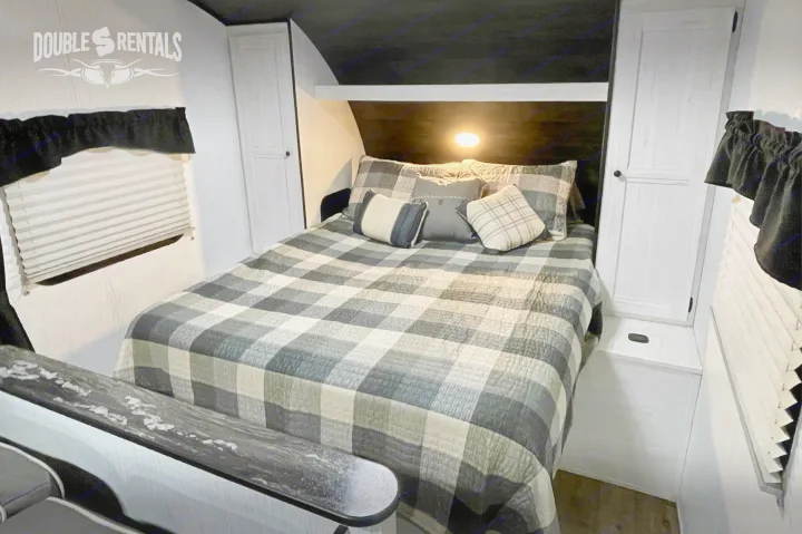 Cozy RV rental bedroom with plaid comforter, warm light, white cabinets, and a pleated window shade. Double S Rentals branding.. Heartland Trail Runner 2021