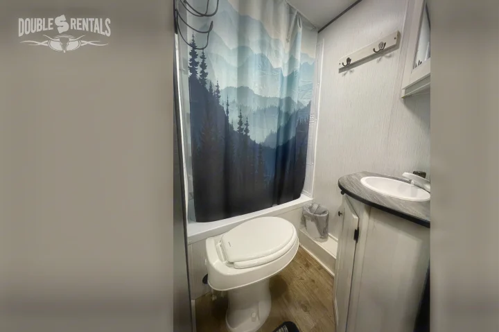 Inside our RV rental: A well-appointed bathroom with a toilet, sink vanity, and a unique mountain-themed shower curtain.. Heartland Trail Runner 2021