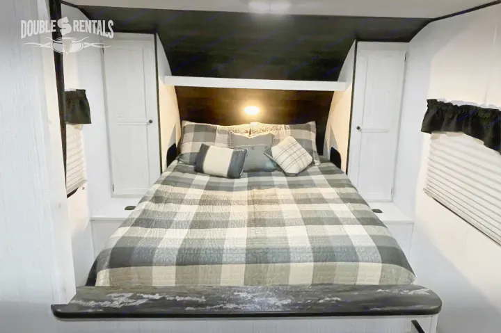 An RV rental bedroom featuring a bed with gray plaid bedding, overhead light, and white built-in storage.. Heartland Trail Runner 2021