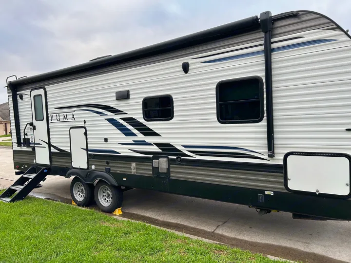 Exterior view of a white Puma travel trailer RV rental, featuring an awning, entry door, steps, and two axles.. Puma Palomino 2021