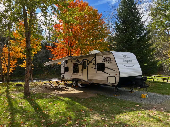Experience an RV rental! A travel trailer is set up at a beautiful fall campsite by a river, surrounded by vibrant autumn trees.. Jayco Jay Flight SLX 2017