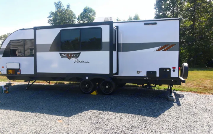 This white travel trailer (RV rental) with an extended slide-out is parked on a gravel lot, ready for adventure.. Forest River Wildwood X-LITE 240BHXL 2024