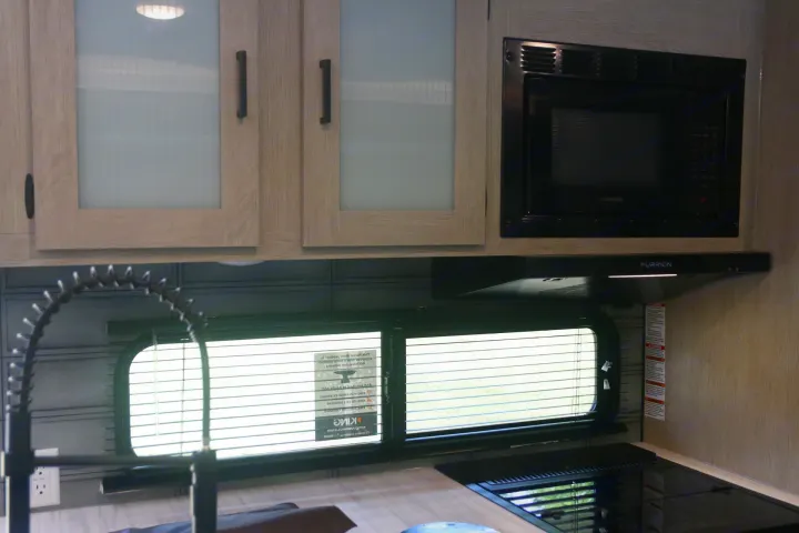 Compact RV rental kitchen with light wood cabinets, Furrion microwave, and cooktop. Includes sink with pull-down faucet.. Forest River Wildwood X-LITE 240BHXL 2024