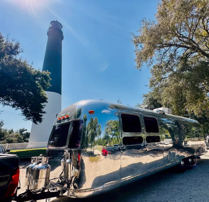 Stunning chrome Airstream RV rental shines by a lighthouse under the sun. Reflecting nature's beauty on your adventure.. Airstream Sovereign 1973