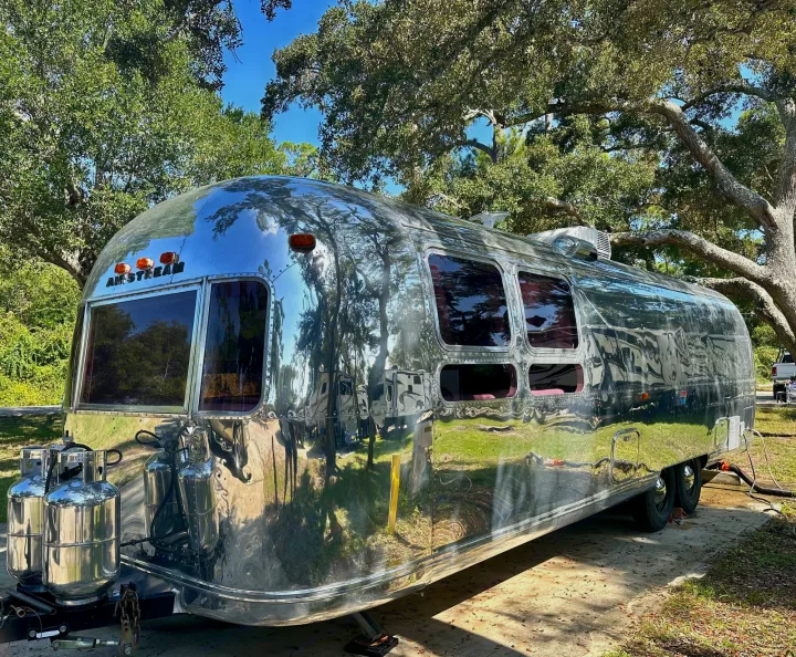 This gleaming Airstream travel trailer, ideal for an RV rental, is parked among trees, reflecting its scenic surroundings.. Airstream Sovereign 1973