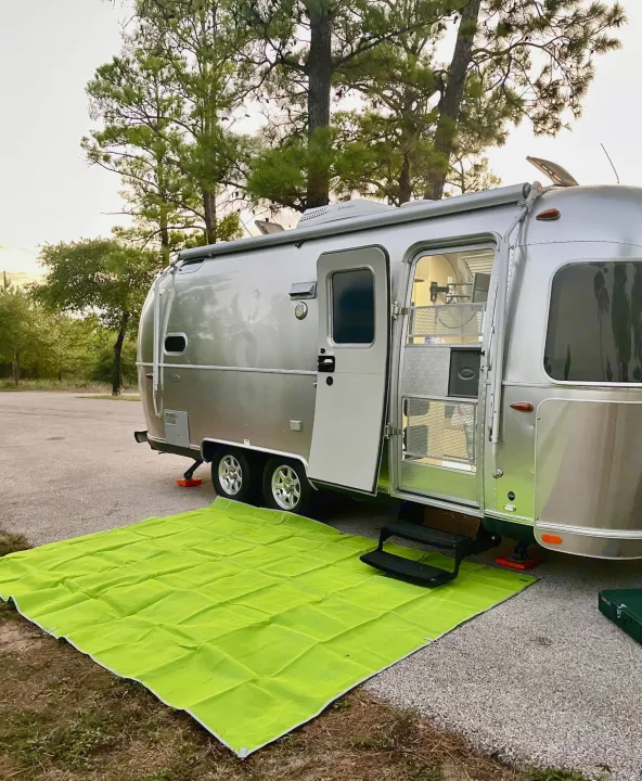 A shiny silver Airstream RV rental is parked at a serene campsite with a vibrant green mat laid out and its door open.. Airstream Flying Cloud 2016