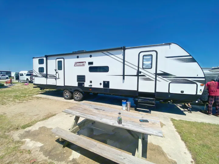 Sunny day at an RV park, featuring a white Coleman travel trailer (RV rental) and a wooden picnic table.. Dutchmen Coleman Light 2022