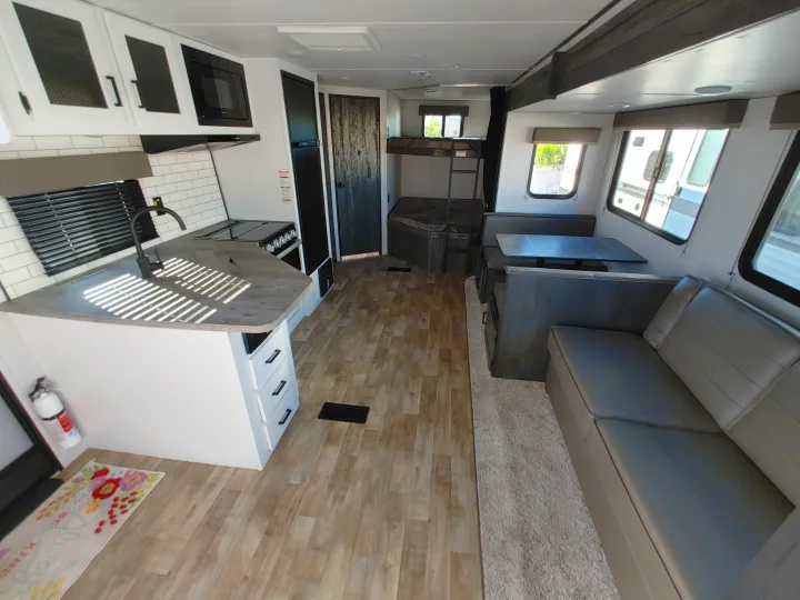 Modern rv rental interior with kitchen, dining booth, and bunk beds, featuring light wood flooring and gray seating.. Keystone RV Hideout 2023