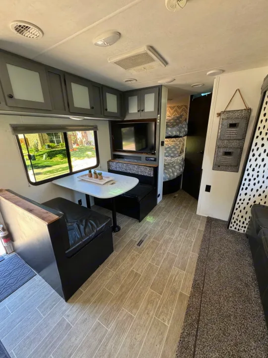 This RV rental interior features a cozy dinette with black seating, overhead cabinets, a TV, and bunk beds. Wood-look floor.. Heartland RVs Trail Runner 2020