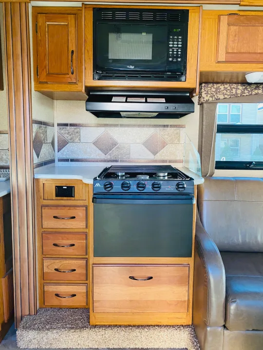 RV rental kitchen featuring a High Pointe microwave, Wedgewood Vision gas oven/stove, countertop, cabinets, and adjacent sofa.. Jayco Redhawk 2016