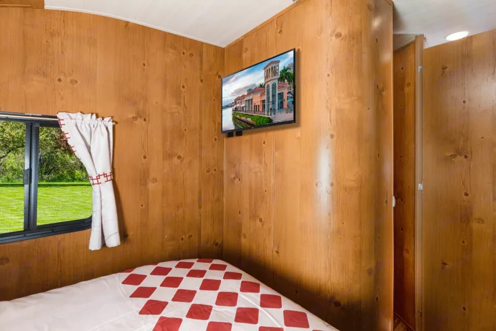 This RV rental offers a cozy wood-paneled bedroom with a comfy bed, TV, and a window providing a green outdoor view.. Gulf Stream Cruiser 2021
