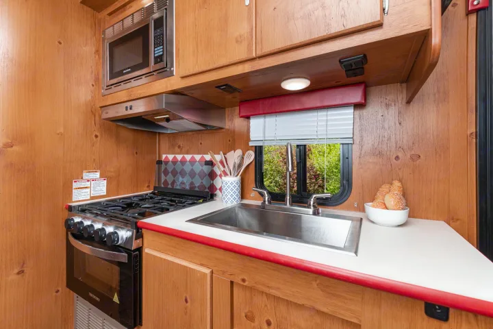 Compact RV rental kitchen features wood paneling, microwave, gas stove, stainless sink, and bright window view.. Gulf Stream Cruiser 2021