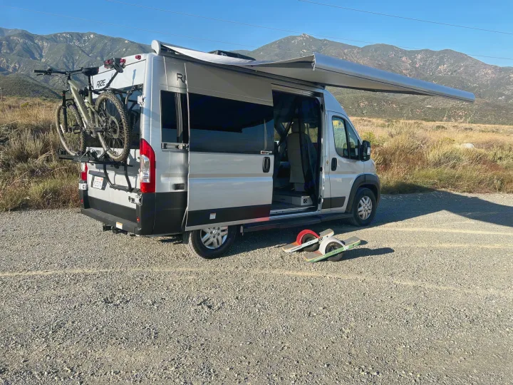 This RV rental camper van with mountain bike & Onewheels, parked on gravel. Ideal for outdoor adventures in scenic mountains.. Thor Motor Coach Other 2023