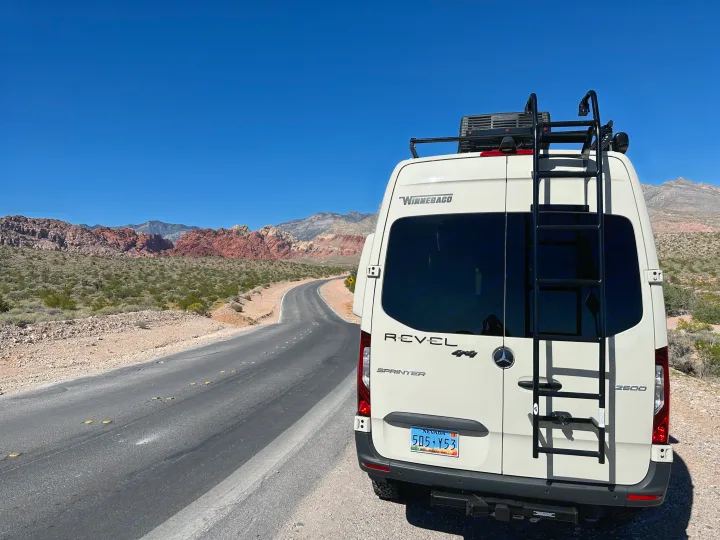 A Winnebago Revel 4x4 rv rental parked on a winding desert road, surrounded by stunning red rock mountains under a clear sky.. Mercedes Revel 2024
