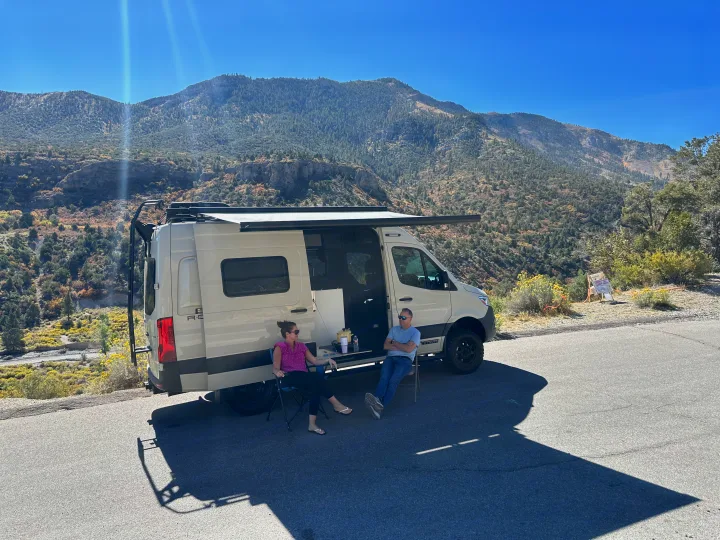 Relaxing outside an **rv rental** with mountain views. A couple enjoys the shade of an awning by their camper van.. Mercedes Revel 2024