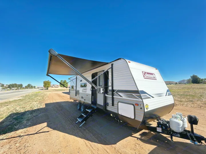 A Coleman travel trailer with an extended awning is parked on a dirt lot under a clear blue sky, ideal for RV rental trips.. Coleman Lantern lt 2023