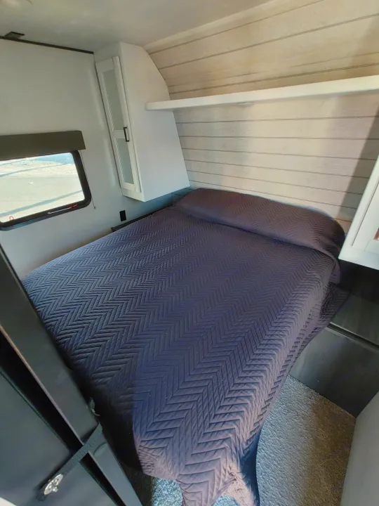 Inside an RV rental bedroom, featuring a comfy bed with a quilted cover, overhead cabinets, and a window.. Keystone RV Hideout 2022