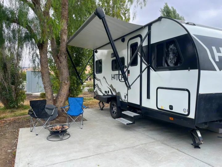 An RV rental site features a white and black travel trailer with an extended awning, two chairs, and a fire pit.. Cruiser RV Hitch 16RD 2022