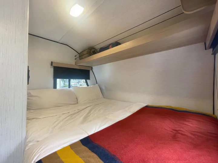 RV rental bedroom with a comfortable bed, white linens & red blanket, overhead shelf, and window.. Cruiser RV Hitch 16RD 2022