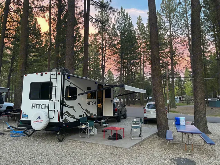 A white RV rental parked at a serene, wooded campsite at sunset, with an outdoor living area.. Cruiser RV Hitch 16RD 2022