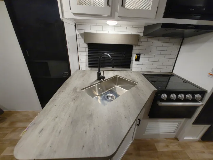 A modern RV rental kitchen featuring a grey countertop, black sink and stove, white subway tile, and overhead cabinets.. Keystone RV Hideout 2023