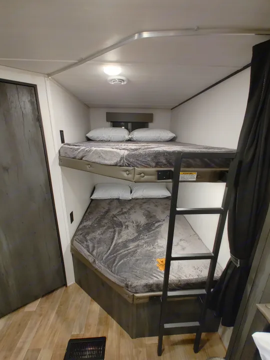 Inside an RV rental, this bunk bed setup offers two cozy sleeping areas with grey bedding and a ladder.. Keystone RV Hideout 2023