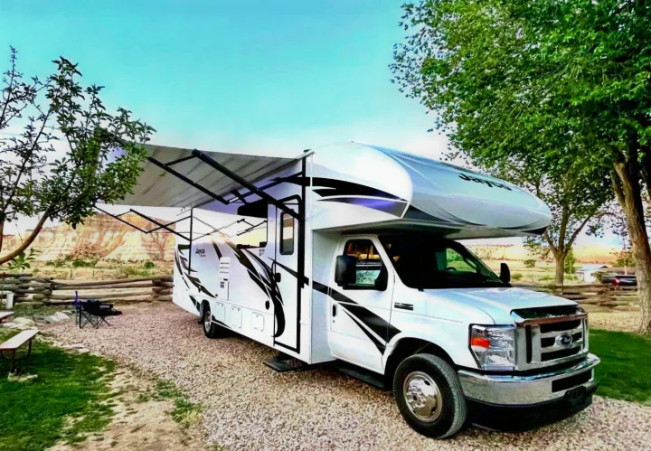 Relaxing RV rental experience! A white Jayco RV with its awning deployed, parked on a gravel site amidst trees with scenic cliffs beyond.. Jayco Redhawk 2022