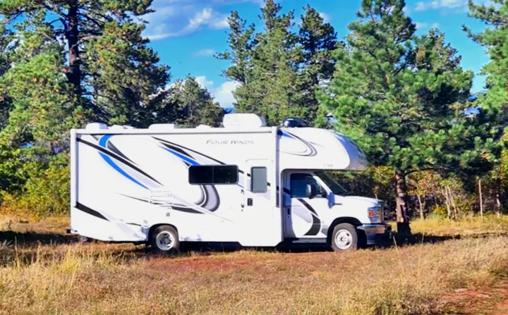 White Four Winds RV rental, a Class C motorhome, nestled among tall pine trees in a sunny clearing. Your camping adventure awaits!. Thor Motor Coach Four Winds 2022