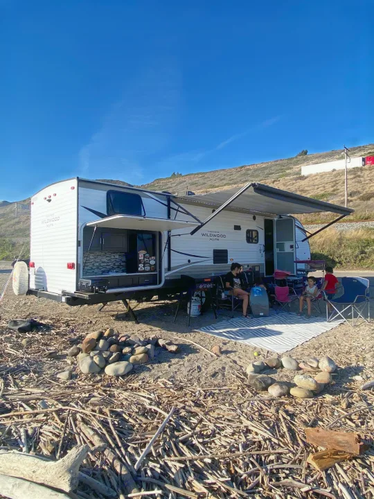 RV rental: Travel trailer with outdoor kitchen for beach camping on driftwood terrain. People relax under awning.. Forest River Wildwood X-Lite 2018