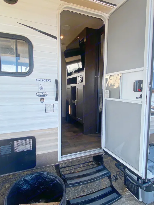 Looking into an RV rental with its main door open, showing the interior. Steps lead up to the entrance, with a trash can nearby.. Forest River Wildwood X-Lite 2018