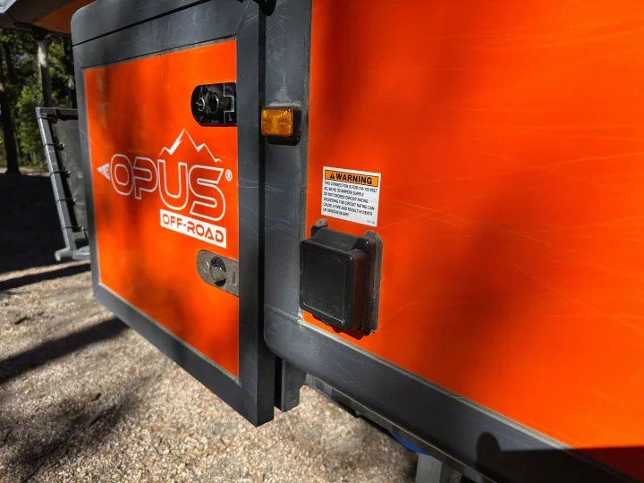 Orange Opus Off-Road trailer side, often used as an RV rental, showing a warning label for its 110-125V power supply connection.. Opus Camper Opus Camper Popup Trailer 2023