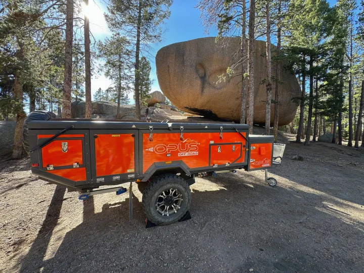 Opus Off-Road camper trailer for RV rental at a scenic boulder-filled campsite amidst tall pines.. Opus Camper Opus Camper Popup Trailer 2023