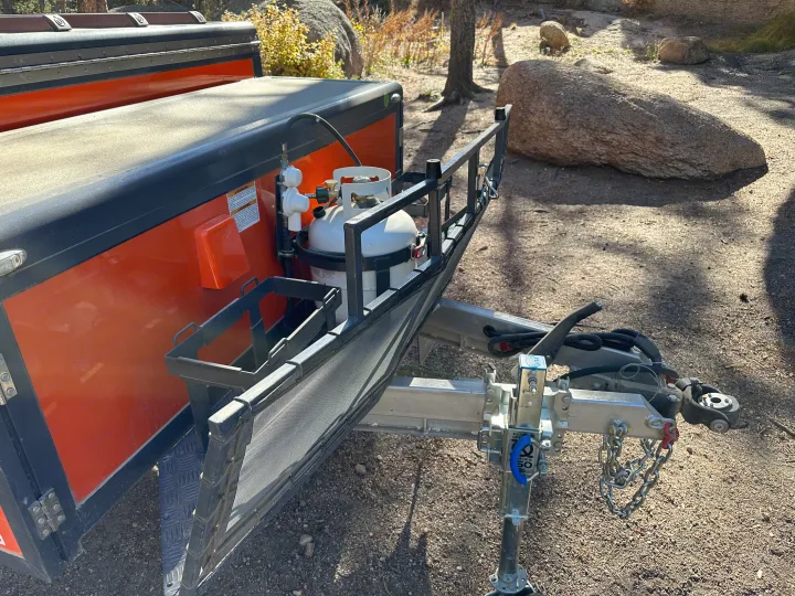 Front of an RV rental-ready overland trailer, featuring dual propane tanks, a mesh rock guard, and robust hitch for outdoor adventures.. Opus Camper Opus Camper Popup Trailer 2023