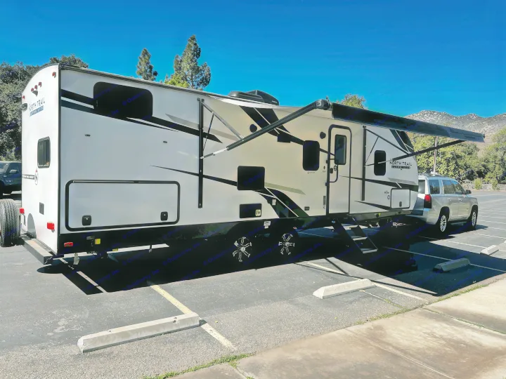 A large white North Trail travel trailer RV rental, with extended awning, parked with an SUV on a sunny day.. Heartland RVs North Trail 2023