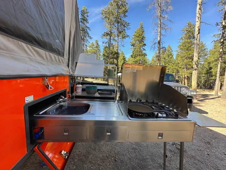 RV rental outdoor kitchen featuring sink, stovetop & storage for camping, nestled among tall pine trees.. Opus Camper Opus Camper Popup Trailer 2023