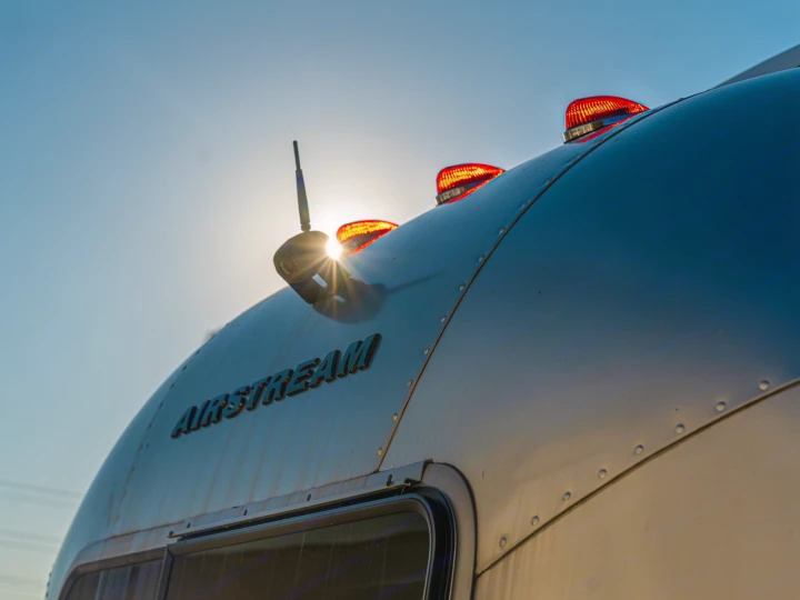 Golden hour Airstream RV rental. Sunburst behind antenna, red marker lights glow on its iconic silver shell.. Airstream Bambi 2023