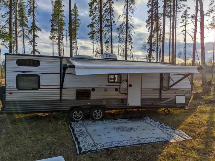 This white and gray travel trailer, likely an RV rental, is set up at a forest campsite with an extended awning and outdoor rug.. Forest River Cherokee Grey Wolf 2014