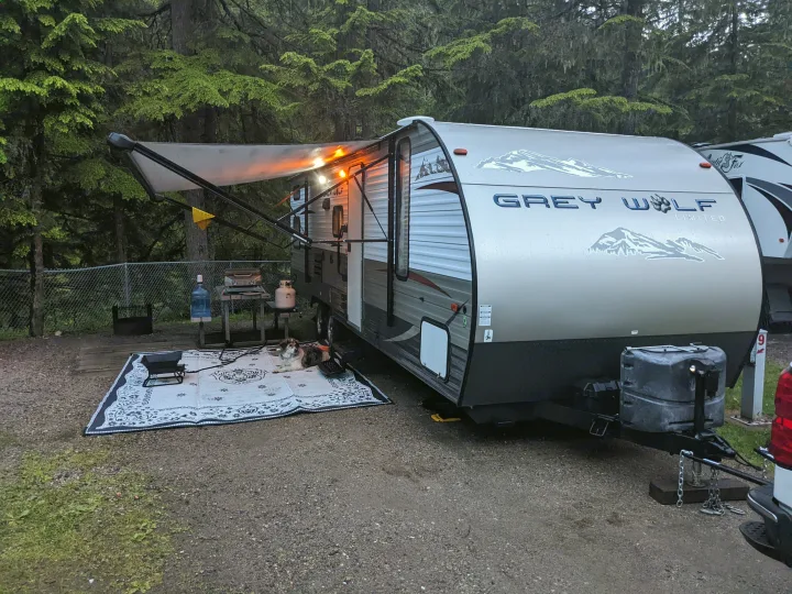 Enjoy a cozy RV rental experience! This travel trailer is parked at a serene campsite with an awning, lights, and a dog.. Forest River Cherokee Grey Wolf 2014