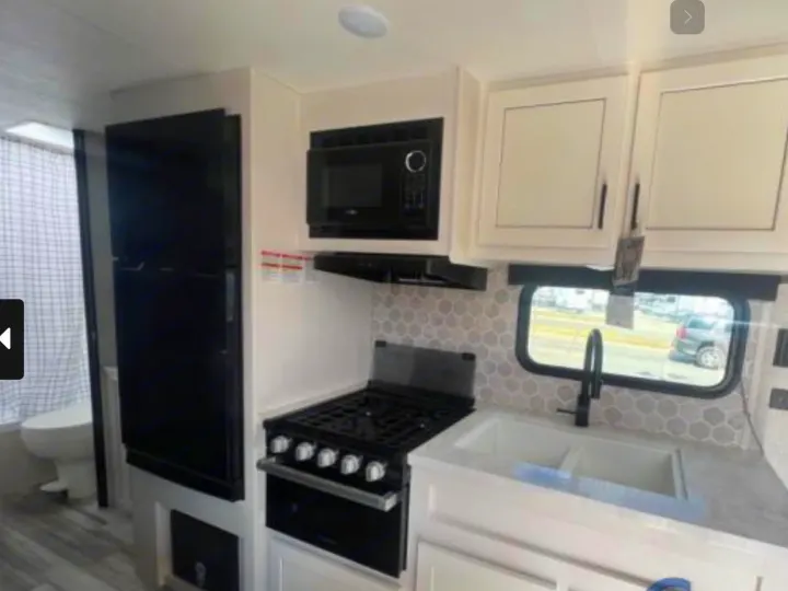 Inside an RV rental: a compact kitchen with microwave, stove, and sink, plus a refrigerator and glimpse of the bathroom.. Jayco Jay Flight 264BH 2024