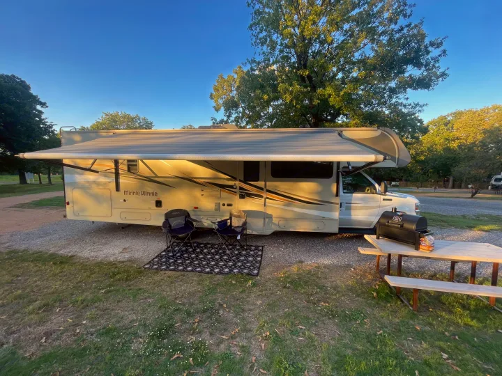 An RV rental campsite view: Extended awning, chairs, rug, and grill on a picnic table under a clear sky.. Winnebago Minnie Winnie 2019