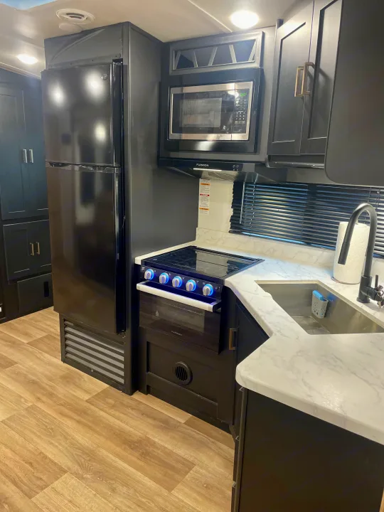 Compact RV rental kitchen featuring black appliances, white marble-look counters, stove, sink, and refrigerator.. Keystone Outback 2022