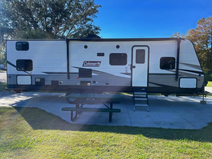 Sunny RV rental setup at a campsite, featuring a white travel trailer, picnic table, and steps. Ready for your adventure!. Dutchmen Coleman Lantern 2019