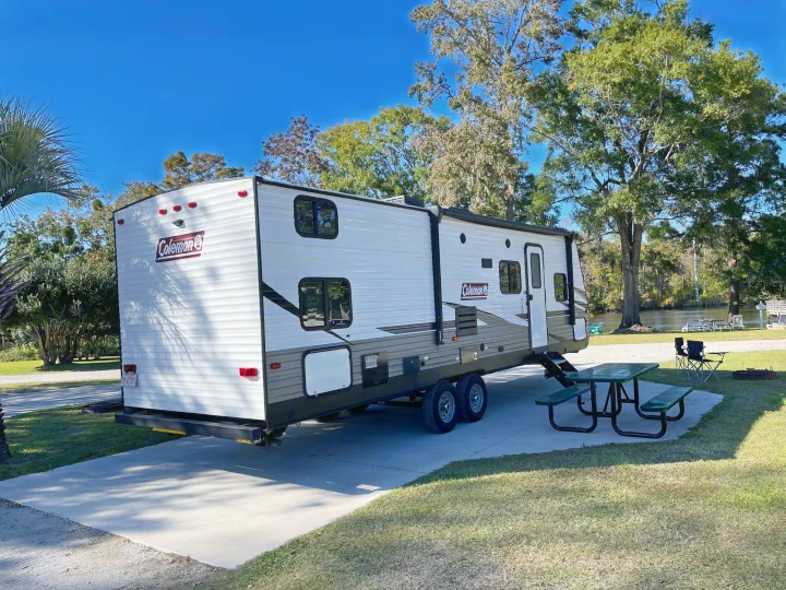 A Coleman travel trailer RV rental at a sunny campground, featuring a picnic area and fire pit by the water.. Dutchmen Coleman Lantern 2019