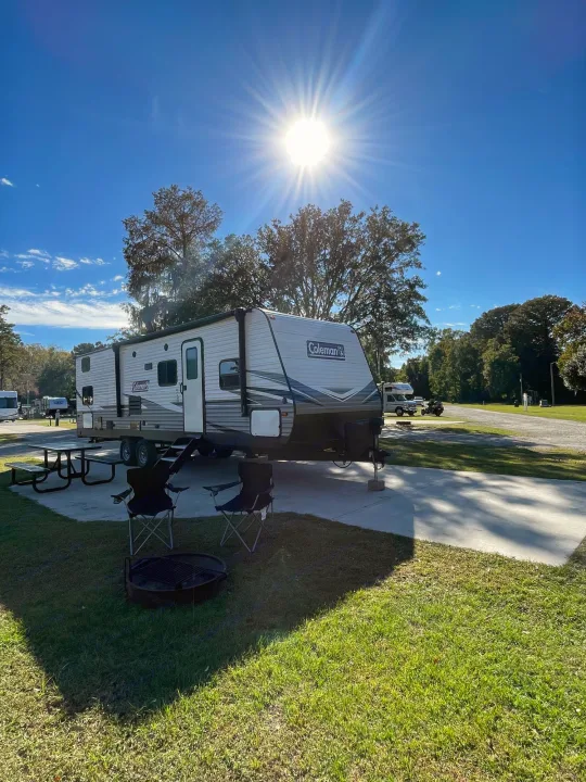 Bright day at an RV rental campsite: trailer, picnic table, chairs, fire pit, and trees under a blue sky.. Dutchmen Coleman Lantern 2019
