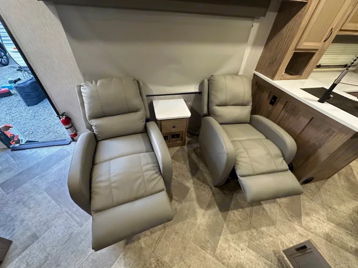 Inside an RV rental, showing two comfy recliners with an end table, and a compact kitchen area to the right.. Palomino Salaire Ultralite 2022