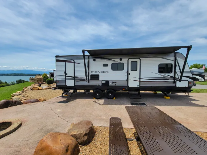 Spacious rv rental parked at a scenic lakeside campsite with a picnic table and mountain views under a blue sky.. Heartland RVs Pioneer 2019