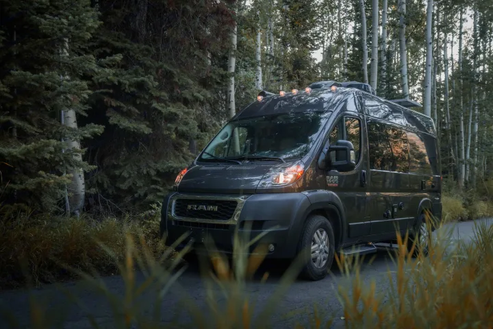 Dark grey RAM ProMaster RV rental van by evergreens & aspens, lights on, parked on a forest road with tall grass.. Thor Rize 2022