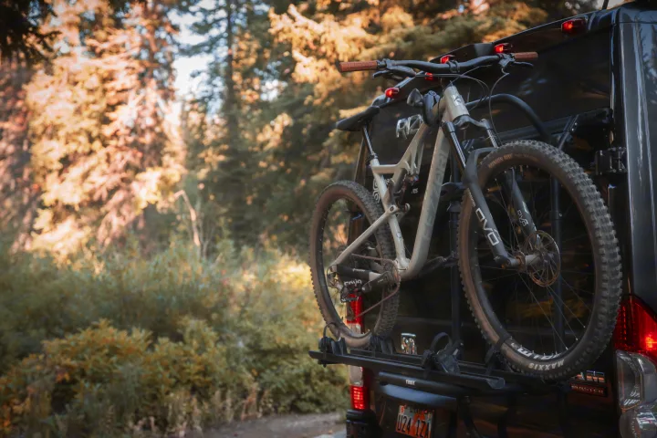 Dark RV rental with two mountain bikes on a rear rack, parked on a dirt road in a sunlit forest.. Thor Rize 2022