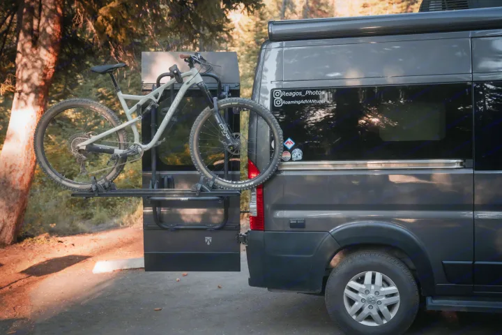 A dark grey RV rental, equipped with a Fezzari mountain bike on a Thule rear rack, is parked amidst a forest setting.. Thor Rize 2022