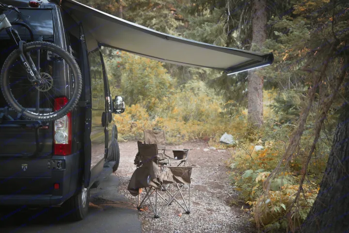 Relaxing rv rental campsite scene: dark camper van with bike, extended awning, and camp chairs among trees.. Thor Rize 2022